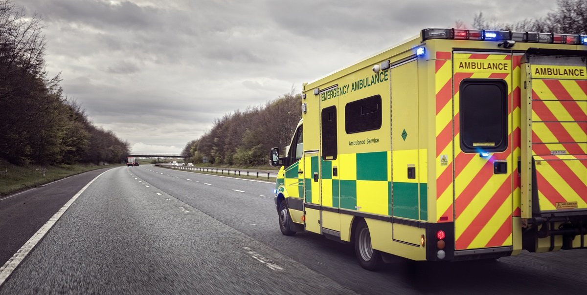 A yellow ambulance with green and yellow checkers on the side drives down a road.